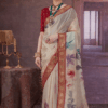 Woman wearing an off-white silk saree with rich red and gold zari border and pastel floral prints, paired with a red blouse and traditional jewellery in a royal indoor setting.