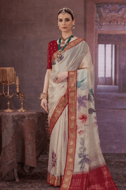 Woman wearing an off-white silk saree with rich red and gold zari border and pastel floral prints, paired with a red blouse and traditional jewellery in a royal indoor setting.